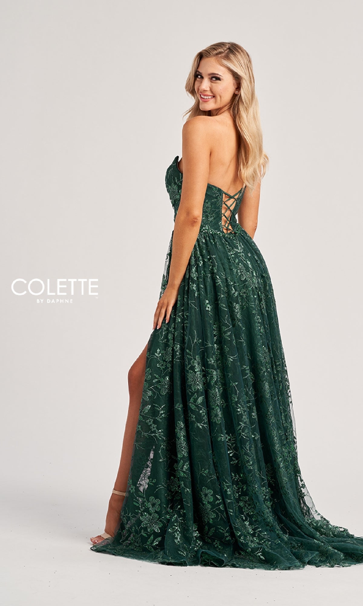 A woman stands sideways, smiling, in the Long Prom Dress: Colette CL8260—a dark green strapless lace gown with a slit and an elegant corset-style lace-up back.